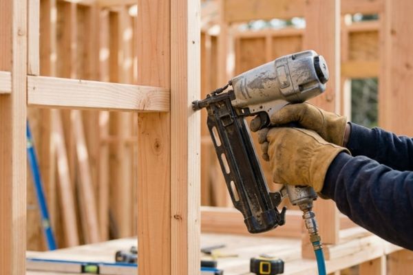 A detailed 1:1 close-up photograph of a carpenter's gloved hands using a pneumatic nailer to secure a wooden stud within the exposed wall framework of a new ADU.